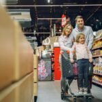 Family enjoying a shopping trip in a supermarket, with a child in a cart and parents smiling.