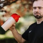 Adult man holds a protein shake in a red lid tumbler outdoors, wearing a black shirt.