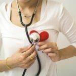Unrecognizable female wearing white shirt while standing on white background with diaphragm of stethoscope on red handmade heart in room